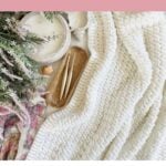 White crochet throw blanket with two crochet hooks on a wooden tray, beside a bowl, greenery, and a pink-patterned cloth on a flat surface. Text reads "Easy Crochet Blanket Pattern.