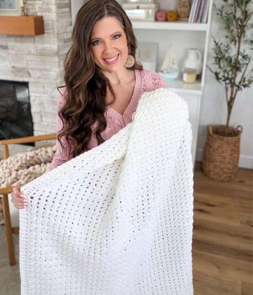 Woman with long brown hair holds and displays a large white honey weave crochet blanket in a cozy, well-lit living room.