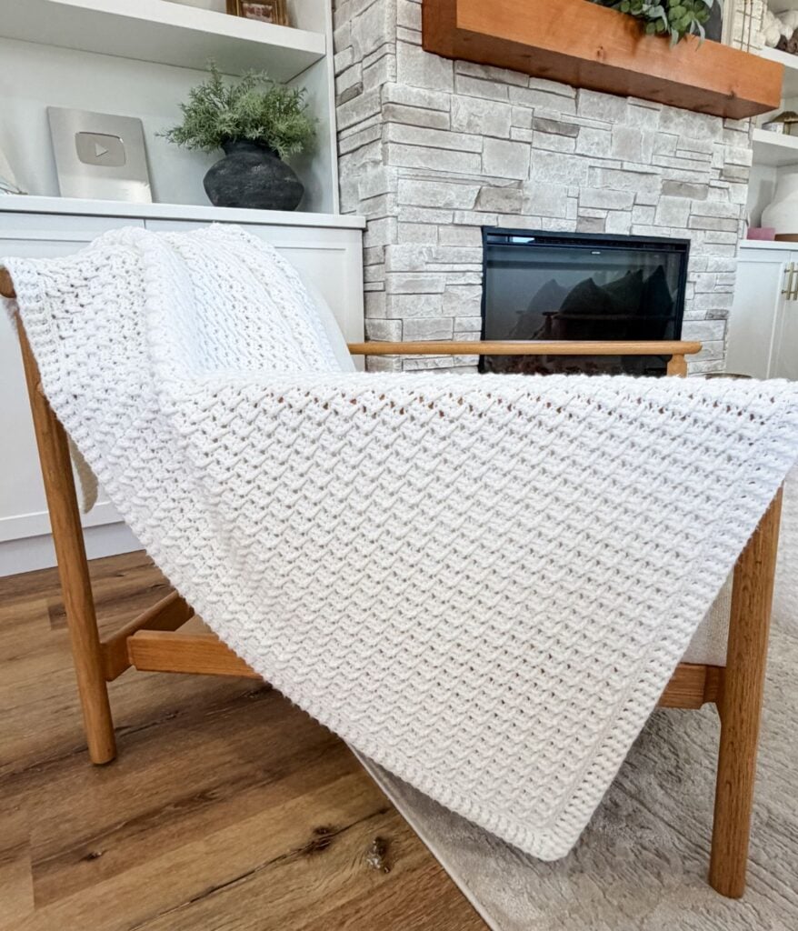 A white honey weave throw blanket is draped over a wooden chair in front of a stone fireplace and built-in shelves.