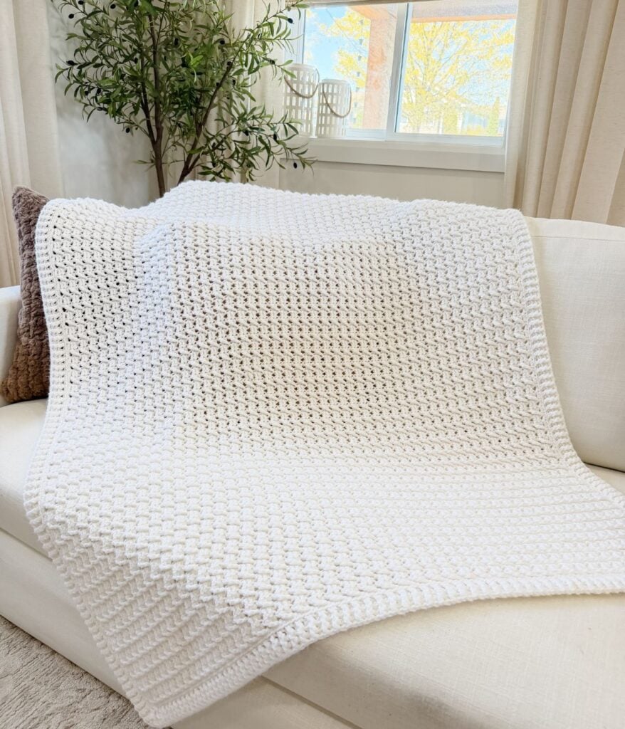 A white Honey Weave knit blanket draped over a light-colored sofa with a brown pillow, near a window and a green potted plant.