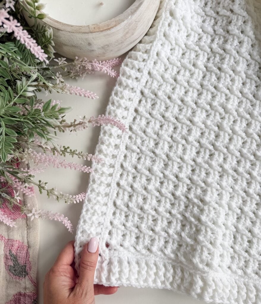 A hand holds the corner of a textured white throw blanket with a subtle crochet pattern near a bowl, accented by pink and green artificial flowers on the side.