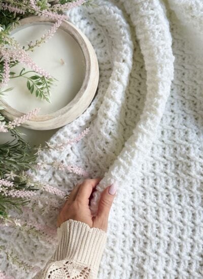A hand holds a textured white Honey Weave throw blanket next to a wooden bowl candle and a floral arrangement with pink flowers and green leaves.