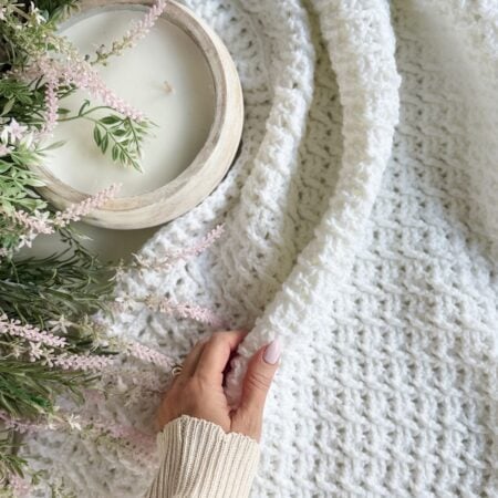 A hand holds a textured white Honey Weave throw blanket next to a wooden bowl candle and a floral arrangement with pink flowers and green leaves.