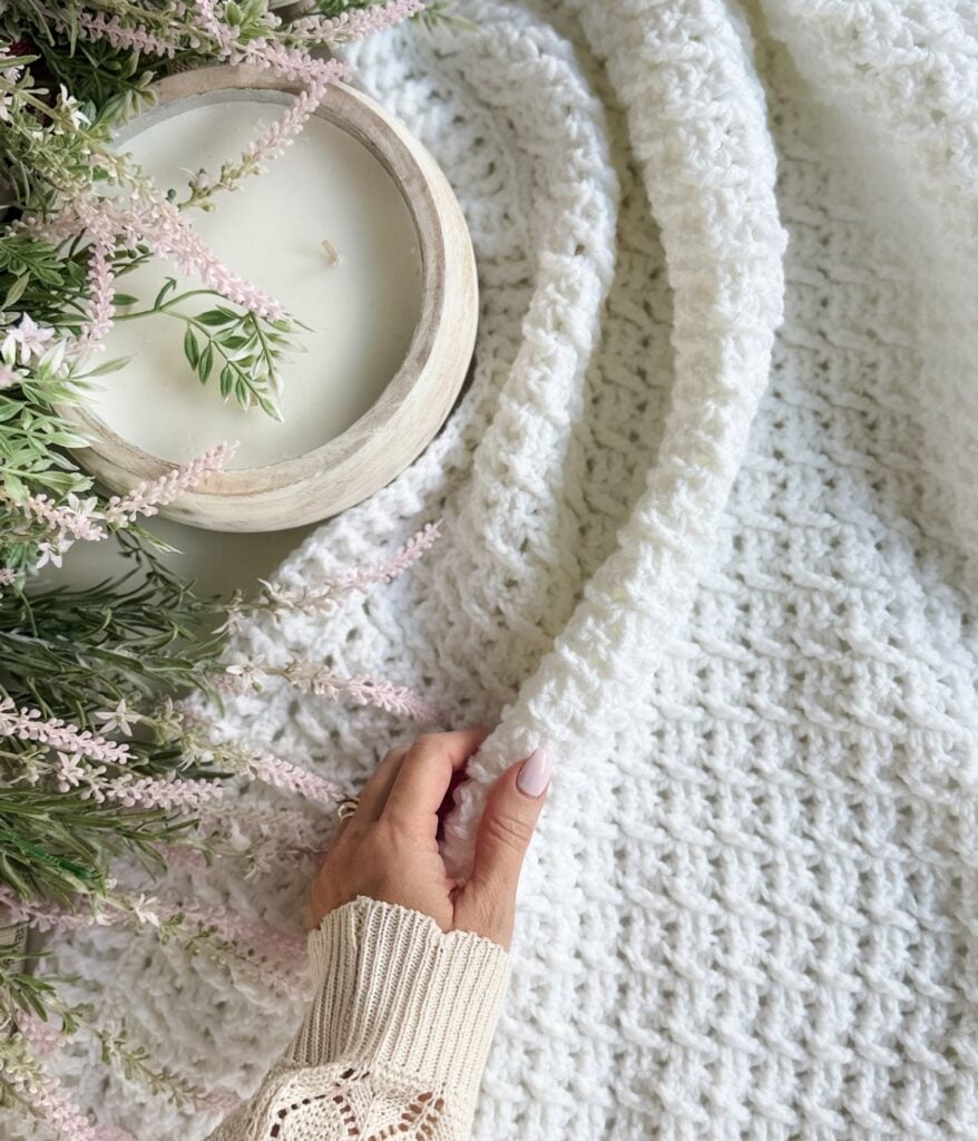 A hand holds a textured white Honey Weave throw blanket next to a wooden bowl candle and a floral arrangement with pink flowers and green leaves.