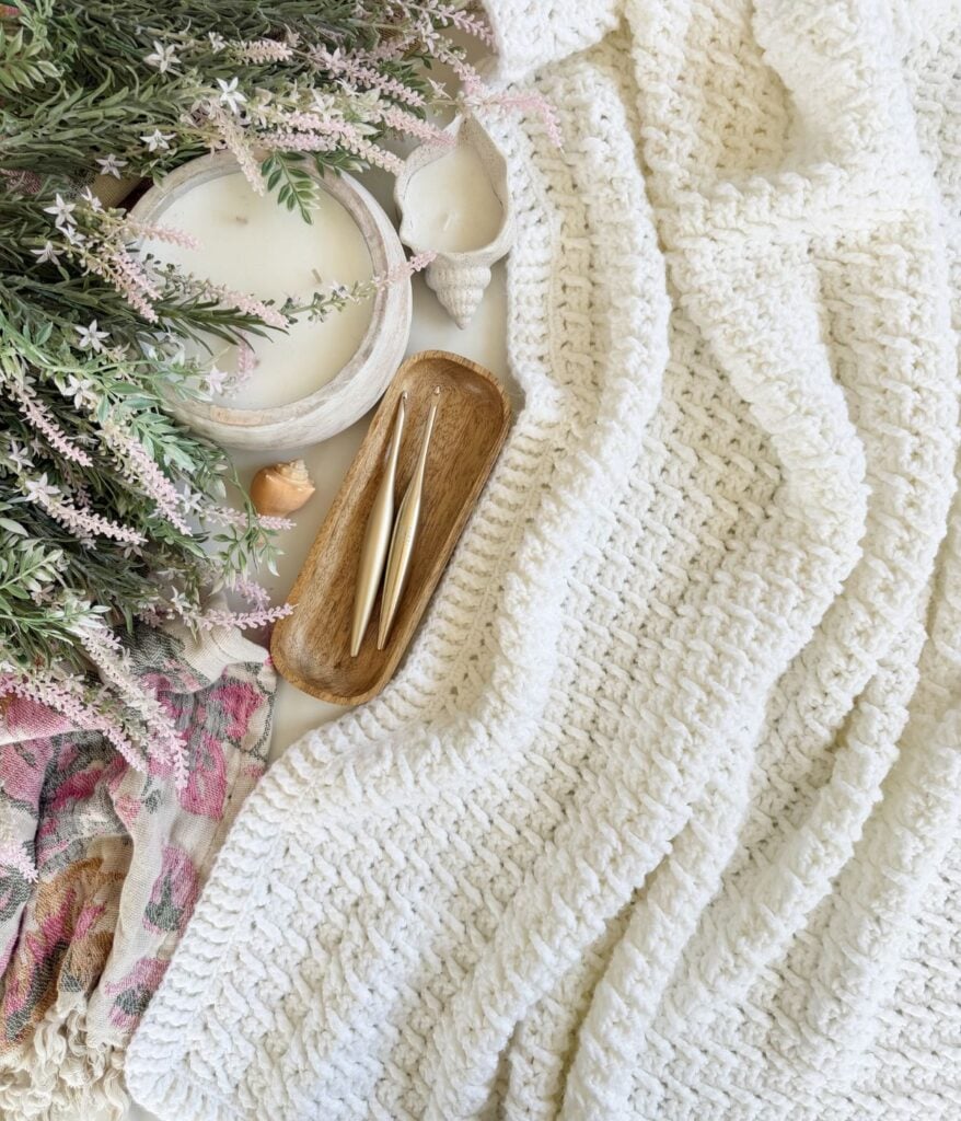 White textured knit throw blanket beside a wooden tray with crochet hooks, a glass candle, greenery, seashells, and a plaid cloth—perfect for showcasing your favorite crochet pattern or adding a touch of cozy Honey Weave style.