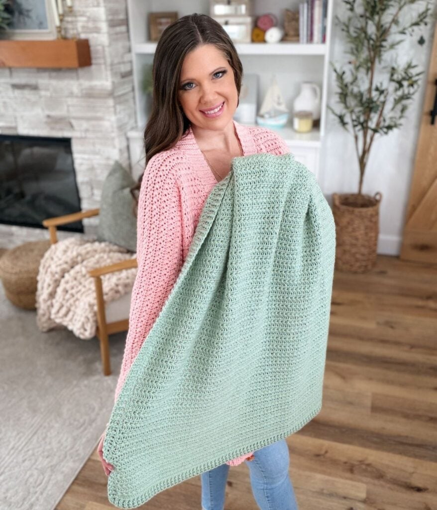 A woman wearing a pink sweater holds a large, light green star stitch blanket in a cozy living room setting.