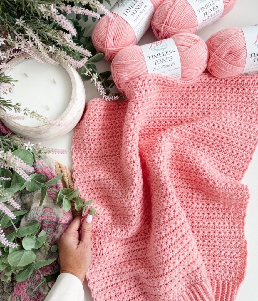 A hand holds a pink textured crochet project—perhaps the start of a Star Stitch Cardigan—next to skeins of pink yarn, a candle, and sprigs of greenery on a white surface.