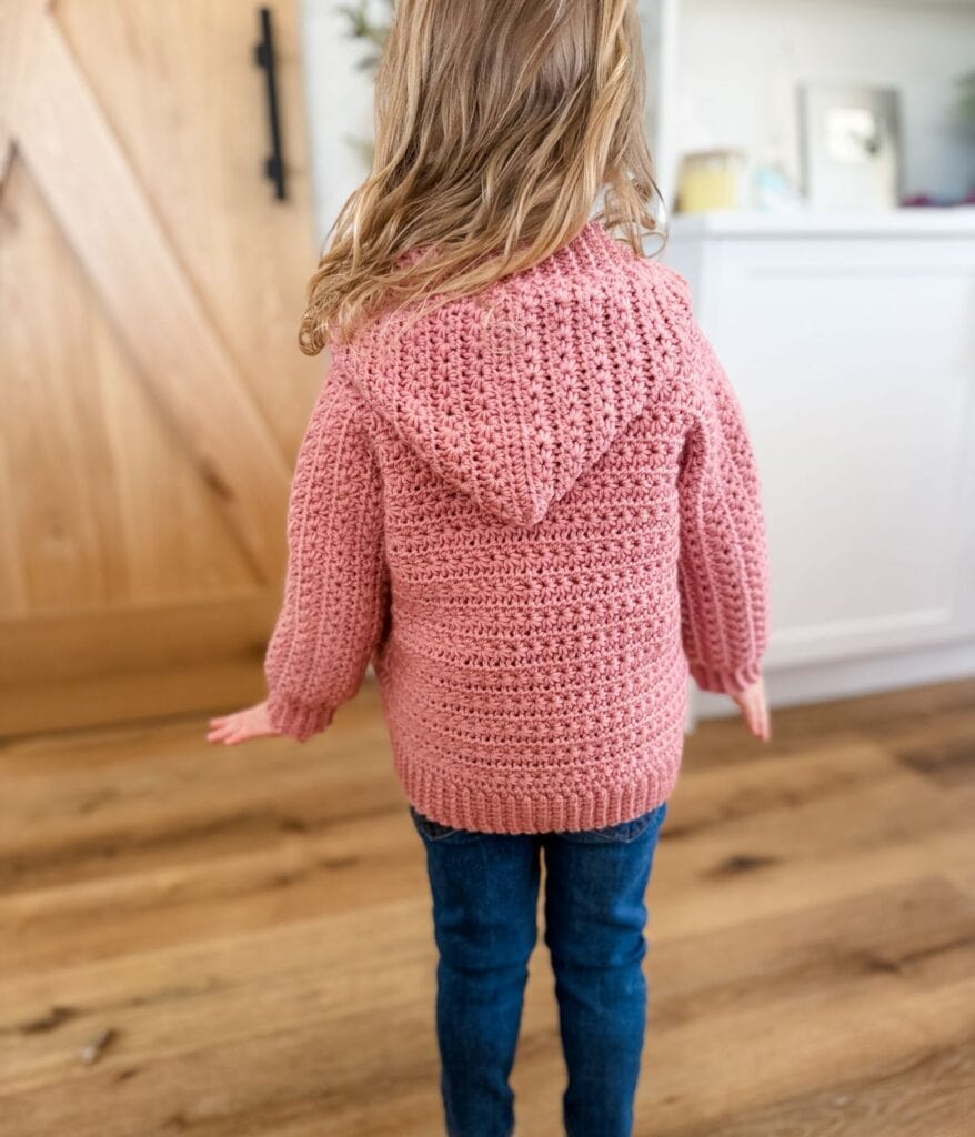 A young child with long hair, seen from behind, wears a pink crocheted cardigan pattern hoodie and blue jeans, standing on a wooden floor in a cozy home setting.
