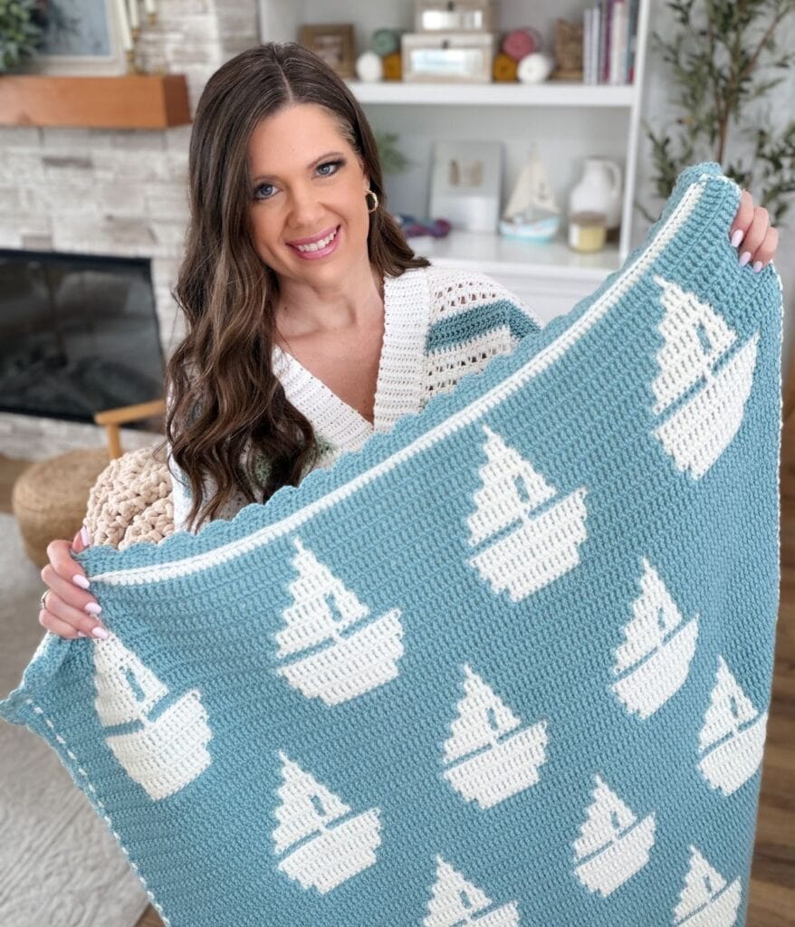 A woman sits indoors holding up a blue mosaic crochet blanket with a white sailboat pattern, showcasing a charming reversible crochet pattern.