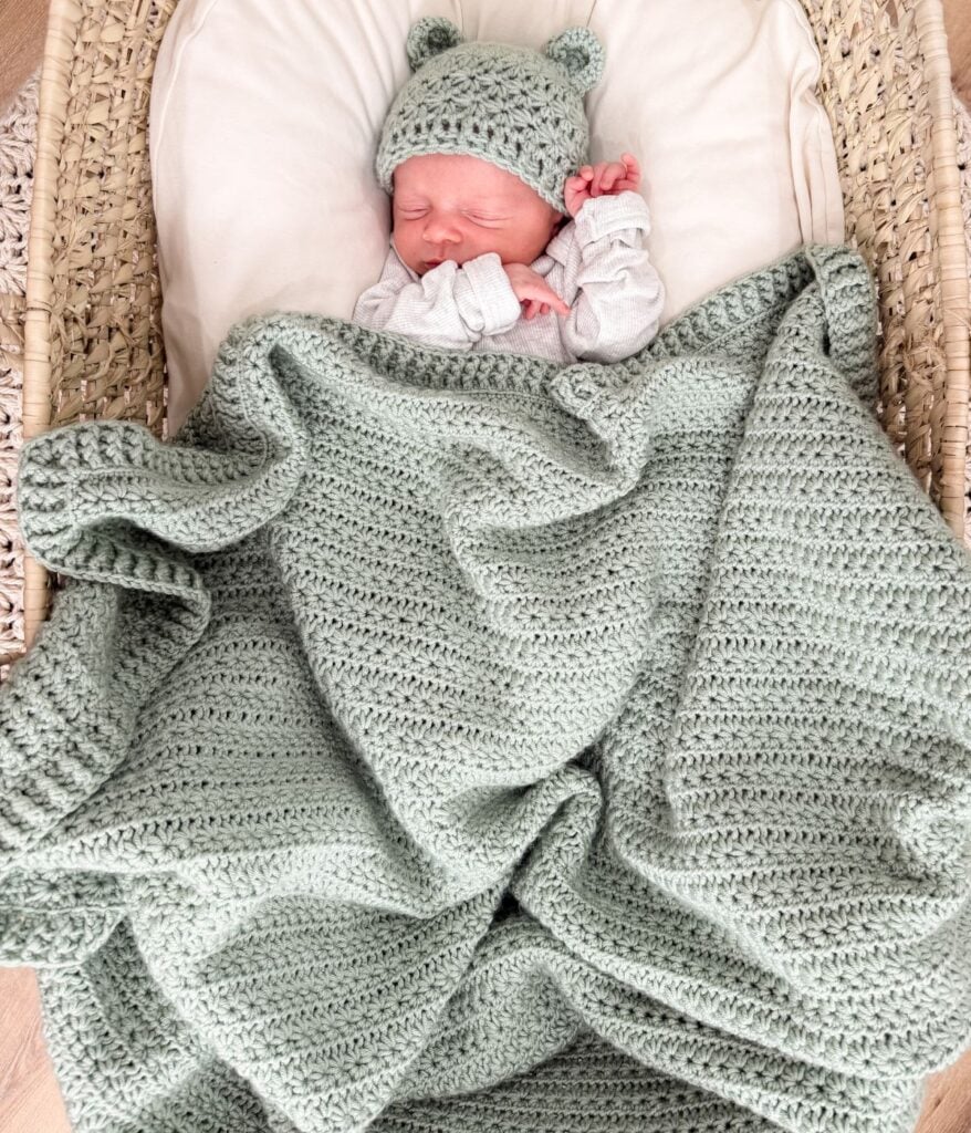 Newborn baby wearing a knitted hat with bear ears sleeps in a basket, cozily covered by a textured sage green star stitch blanket.