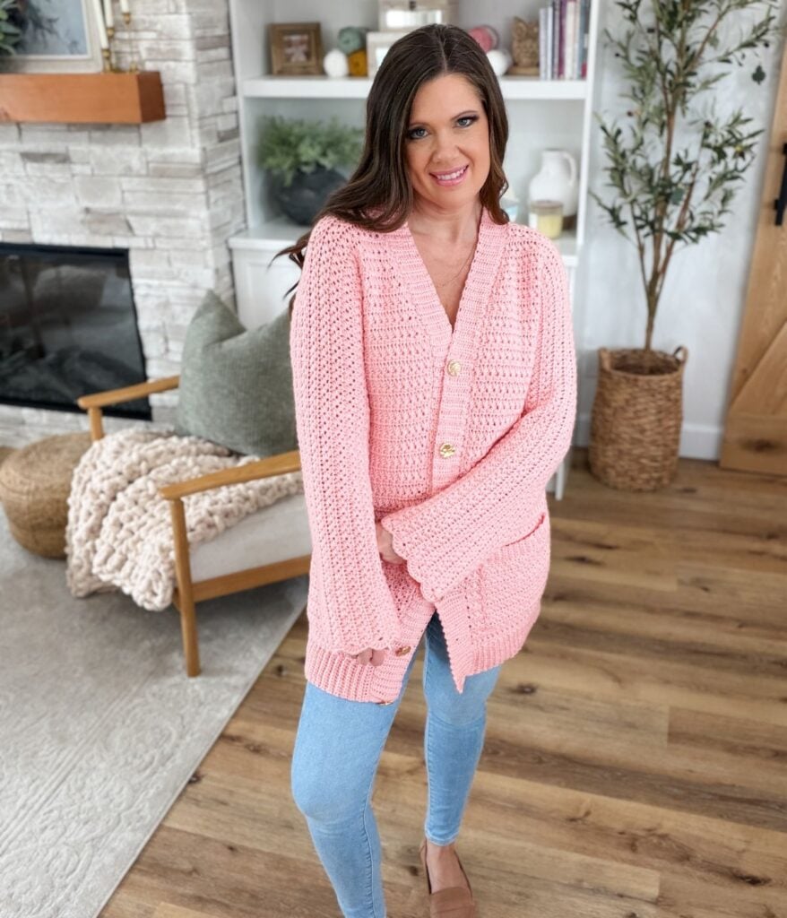 A woman with long brown hair stands indoors wearing a pink crochet cardigan made with a star stitch pattern, blue jeans, and beige shoes. She is smiling and has one hand in her pocket.