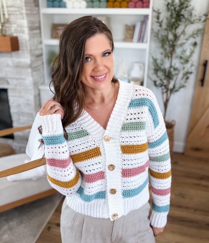 A woman with long brown hair wears MJ's Signature Cardigan, a white knit with colorful horizontal stripes, paired with beige pants, standing indoors in a cozy, modern living space.