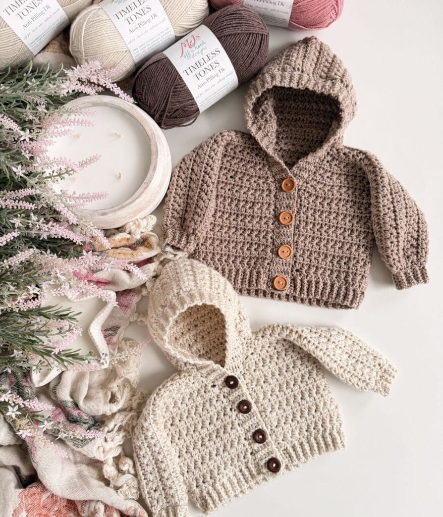 Two crocheted baby hooded sweaters, one beige and one cream, each with buttons, are displayed flat beside yarn skeins, a textured cloth, and a white candle with decorative greenery—perfect for showcasing your favorite cardigan pattern.