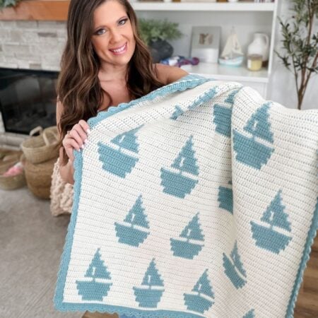 A woman indoors holds a Mosaic Crochet Blanket featuring a repeating pattern of blue sailboats on a white background with a blue border.