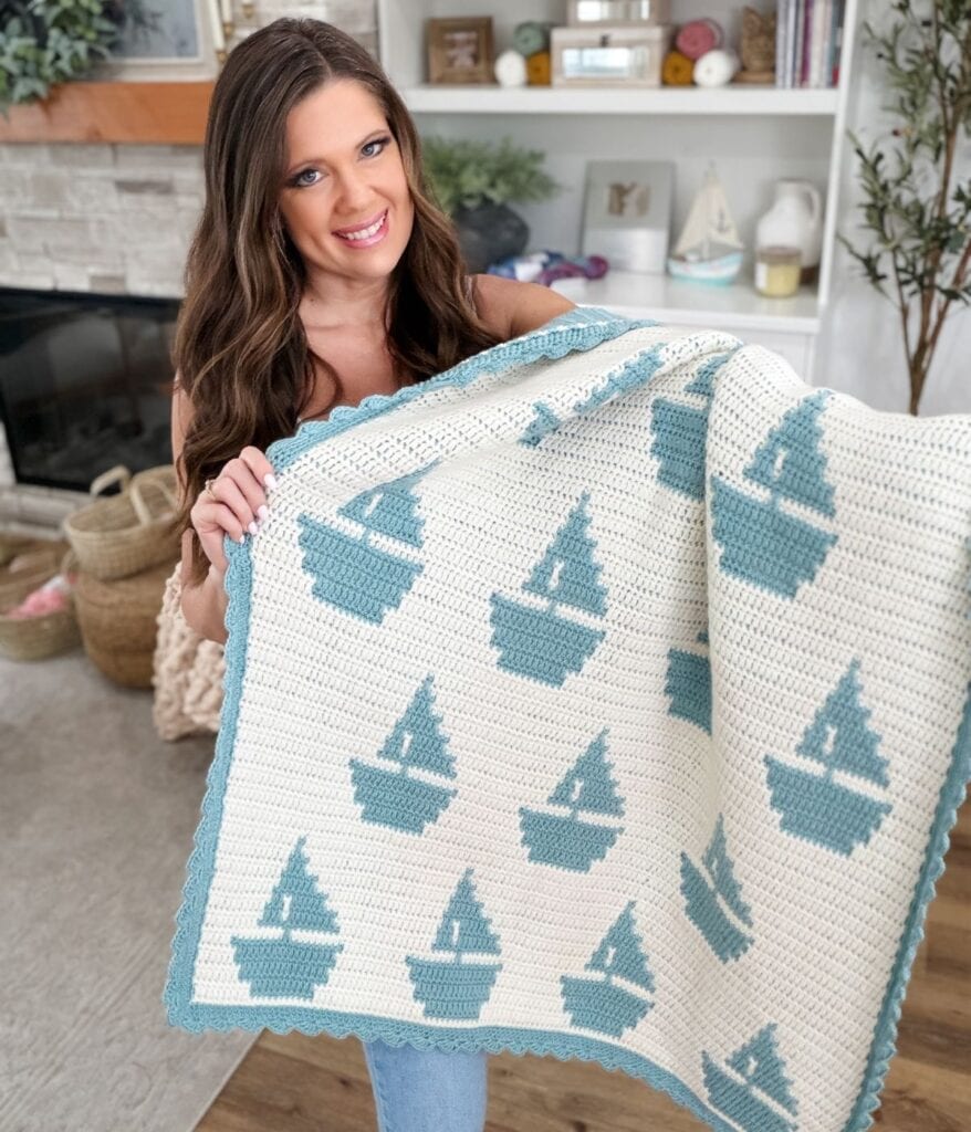 A woman indoors holds a Mosaic Crochet Blanket featuring a repeating pattern of blue sailboats on a white background with a blue border.