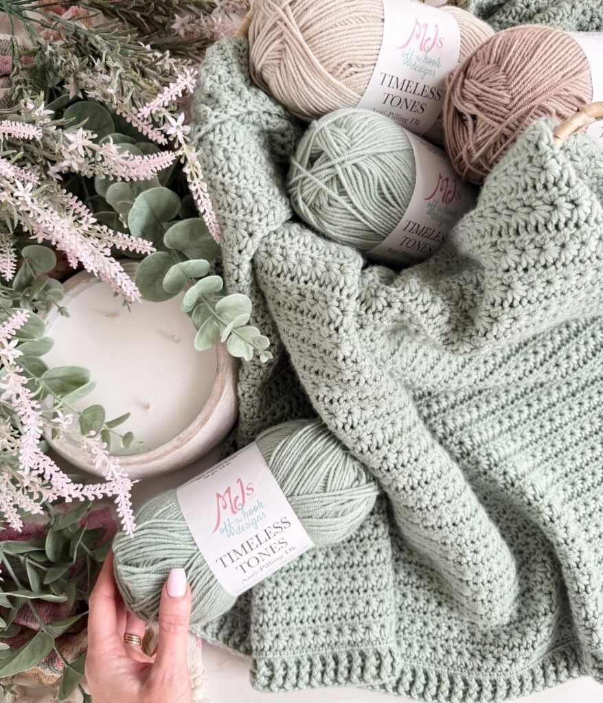 Yarn balls and a crocheted green star stitch blanket are displayed next to a potted plant. A hand holds one of the yarns labeled "Timeless Tones.