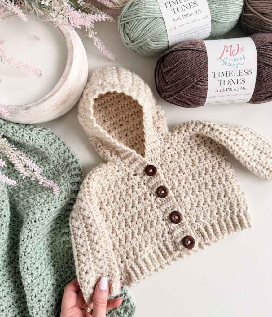 A hand holds a crocheted cream-colored baby hoodie with brown buttons, crafted using a cozy cardigan pattern, placed beside skeins of yarn and a green crocheted blanket on a light surface.