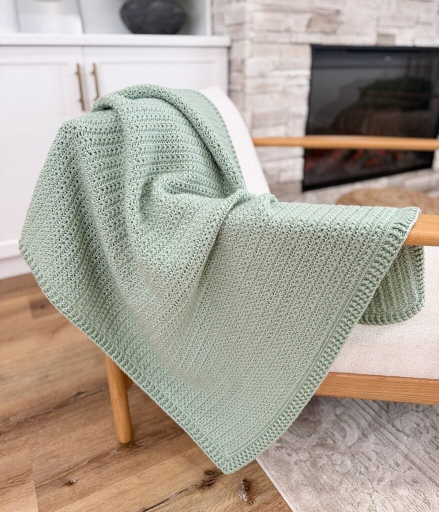 A light green star stitch blanket is draped over the arm of a cream-colored chair in a cozy living room setting.