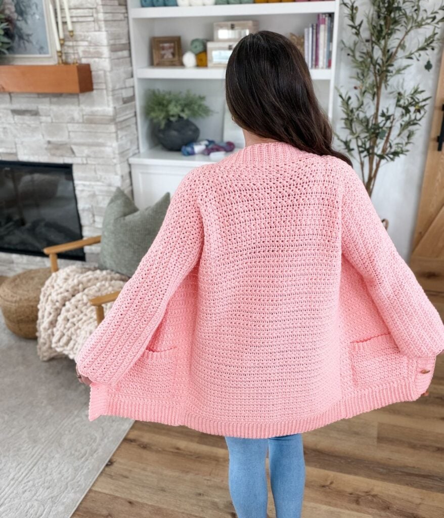 A person with long brown hair wears a pink Star Stitch Cardigan, standing indoors with their back to the camera to highlight the crochet cardigan pattern’s unique texture and shape.