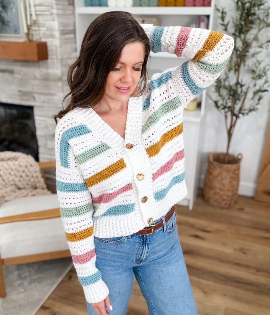 Woman in a colorful striped MJ's Signature Cardigan and blue jeans stands indoors, with one hand touching her hair, in a cozy, modern living room. Find the free crochet pattern to create this vibrant look yourself.