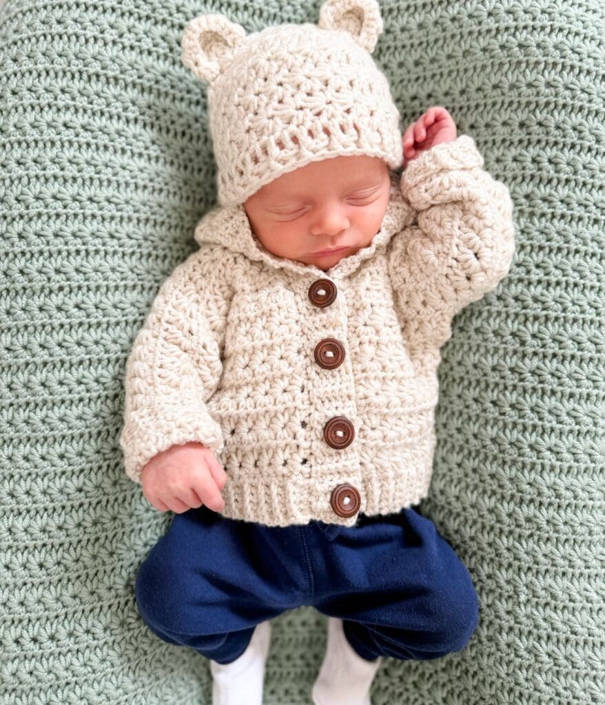 Newborn baby wearing a cream-colored crocheted cardigan pattern with brown buttons and a matching hat with bear ears, lying on a green crocheted blanket.