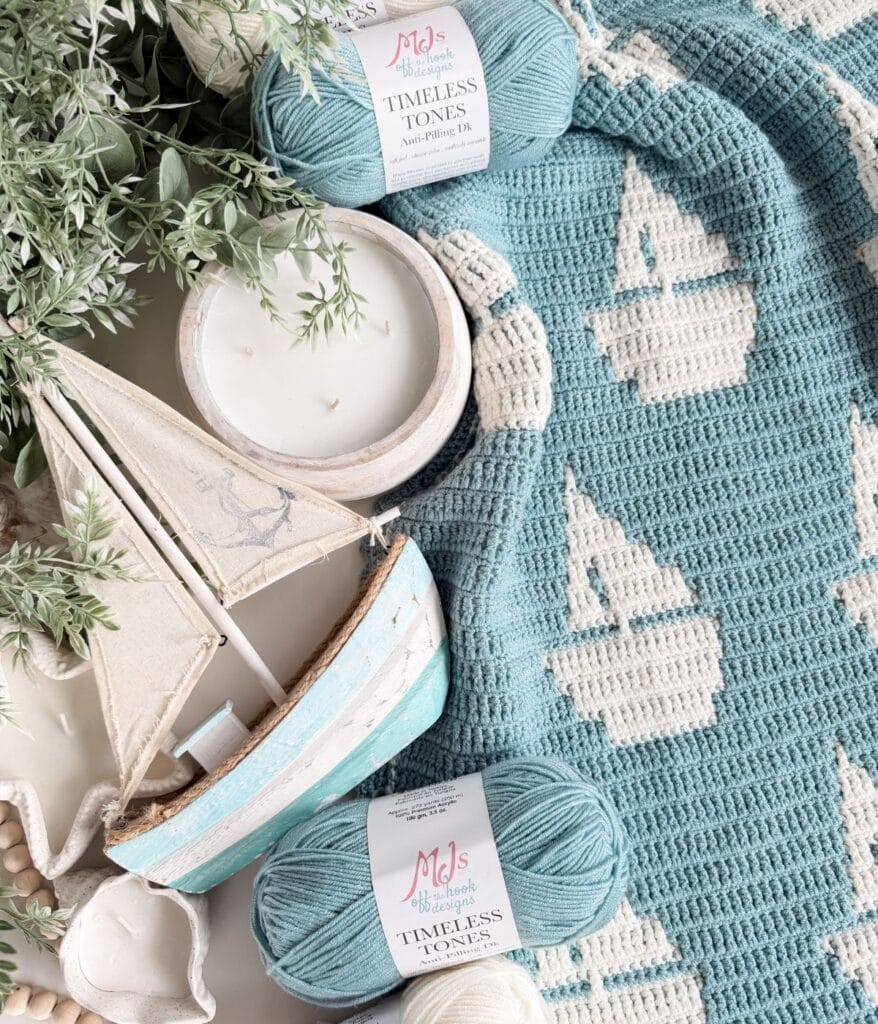 A crocheted blue and white sailboat baby blanket with mosaic crochet blanket patterns, two skeins of blue yarn, a model sailboat, a white candle, and green foliage arranged on a table.