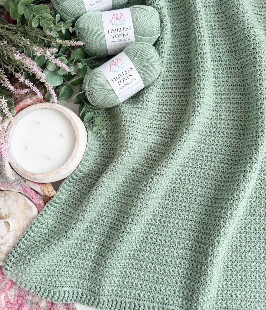 A sage green star stitch blanket is displayed next to three matching yarn skeins, a white candle, and some greenery on a light surface.