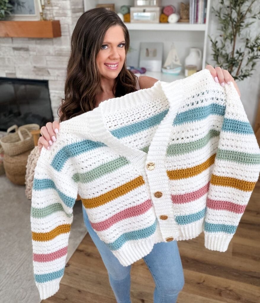 Woman holding up a Signature Cardigan with horizontal pastel stripes in blue, green, mustard, and pink, featuring large buttons—crafted from a cozy Free Crochet Pattern—standing in a warm living room.