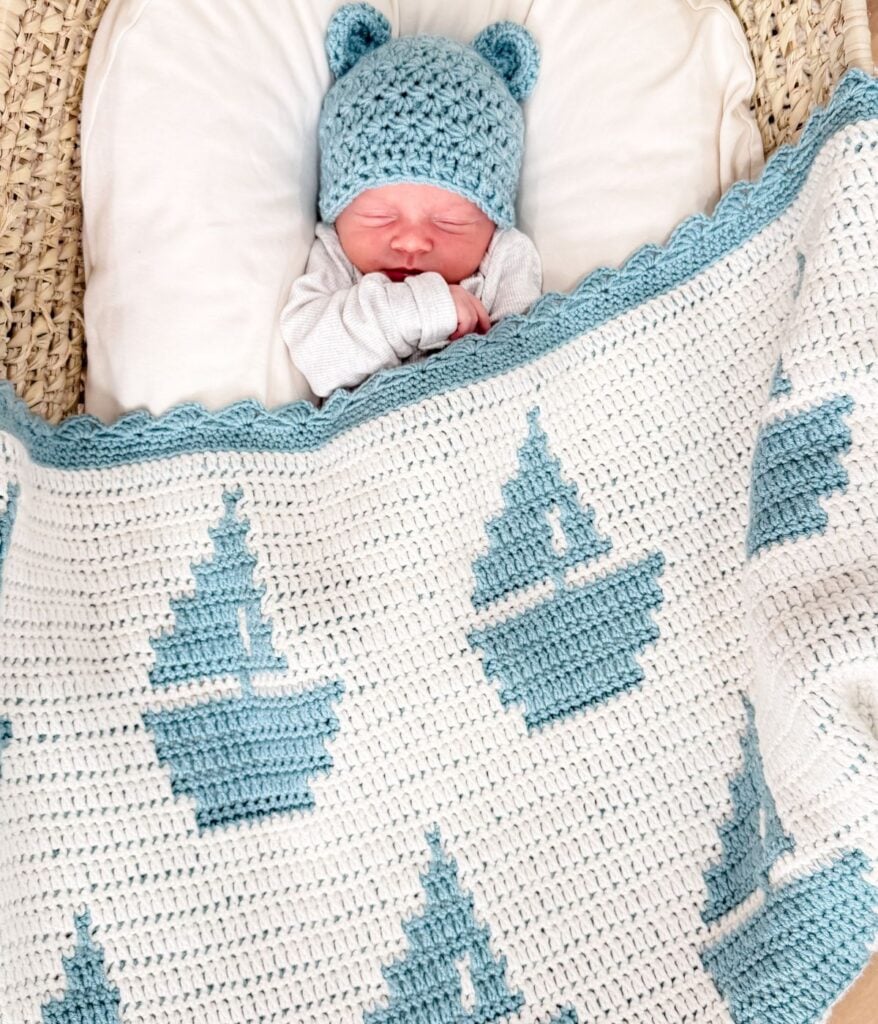 A newborn baby wearing a blue knit hat with bear ears sleeps under a cozy sailboat baby blanket, featuring a reversible crochet pattern in white and blue.