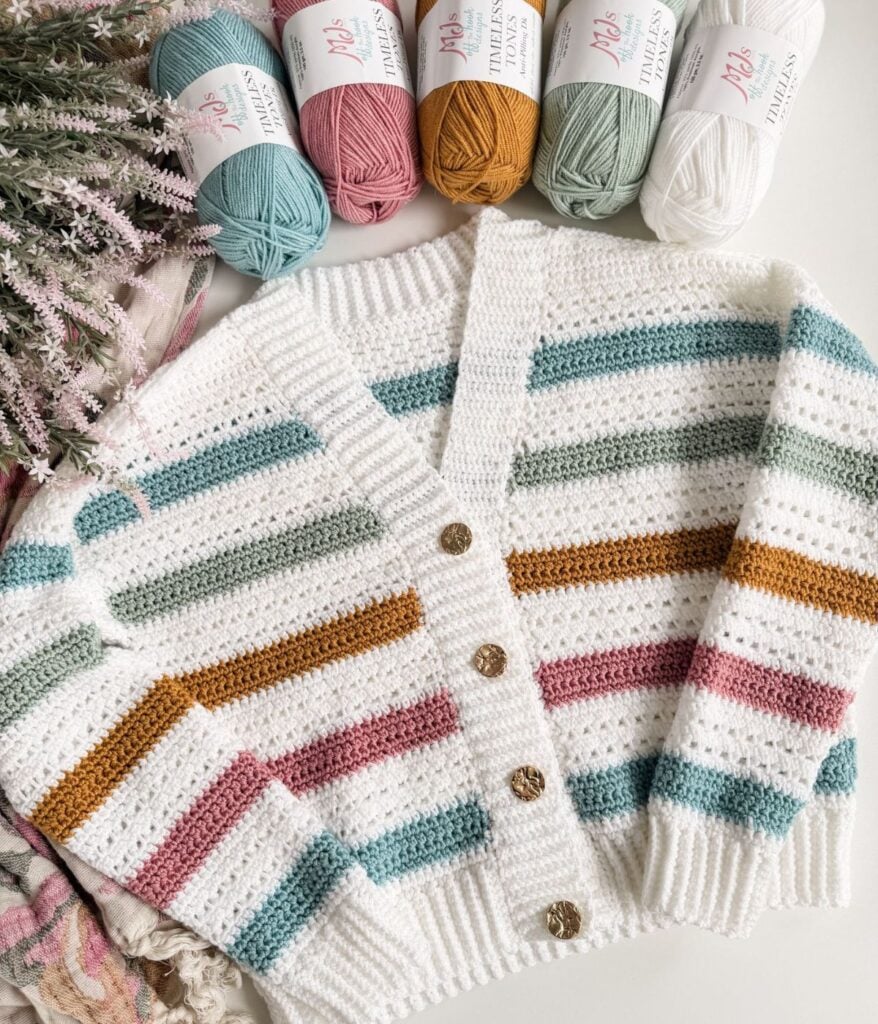 A crocheted cardigan with horizontal stripes in teal, mustard, pink, and white is laid flat with skeins of matching yarn and some dried flowers beside it—perfect for making with MJ's Signature Cardigan free crochet pattern.
