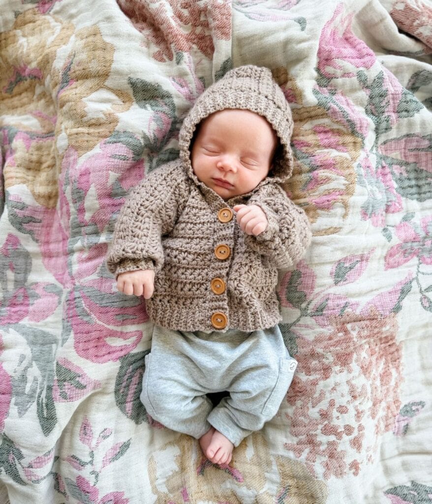 A sleeping baby wearing a knitted brown hoodie with a charming cardigan pattern and grey pants lies on a floral-patterned blanket.
