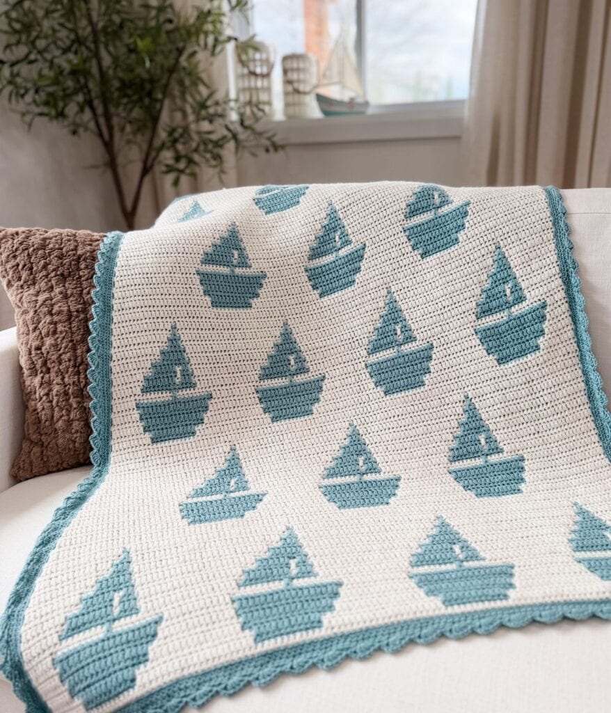 A reversible crochet pattern sailboat baby blanket with blue sailboat motifs and a blue border is draped over a white couch with a brown textured pillow in a bright room.
