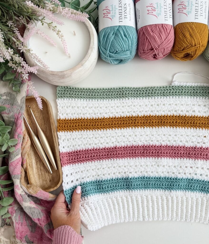 A hand holds a crocheted striped fabric in white, mustard, and teal—perfect for crafting MJ's Signature Cardigan. Nearby are yarn balls, crochet hooks, and a candle. Discover your next project with this free crochet cardigan pattern.