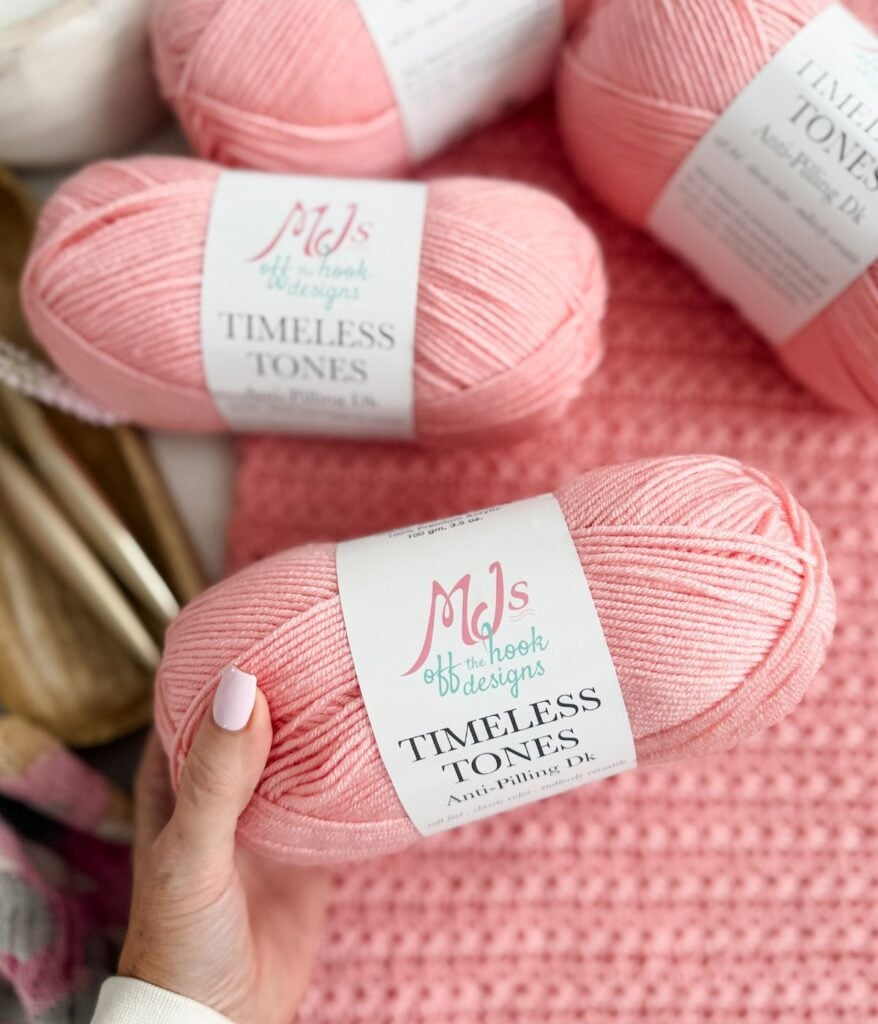 A hand holds a pink skein of "Timeless Tones" yarn by MJ's Off The Hook Designs—perfect for creating a cozy Crochet Cardigan; more skeins and a pink crocheted piece are in the background.