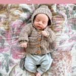 Newborn baby wearing a brown crocheted cardigan with a hood, lying on a floral-patterned blanket. Text above advertises a star stitch crochet cardigan pattern, perfect to pair with your favorite Mosaic Crochet Blanket for a cozy nursery set.