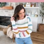 Woman wearing a colorful striped crochet sweater stands in a cozy living room, promoting a spring crochet sweater pattern from MJ's Off the Hook Designs, alongside her latest mosaic crochet blanket creation.