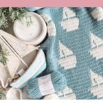 Blue and white crochet blanket featuring a sailboat mosaic baby blanket design, displayed beside a decorative sailboat, greenery, and matching yarn skein. Text highlights this reversible mosaic crochet blanket pattern.