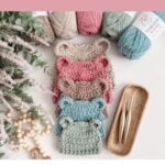 Three crocheted baby hats with bear ears in pink, beige, and blue are displayed with yarn, crochet hooks, wooden beads, flowers, and a mosaic crochet blanket pattern on a light surface.