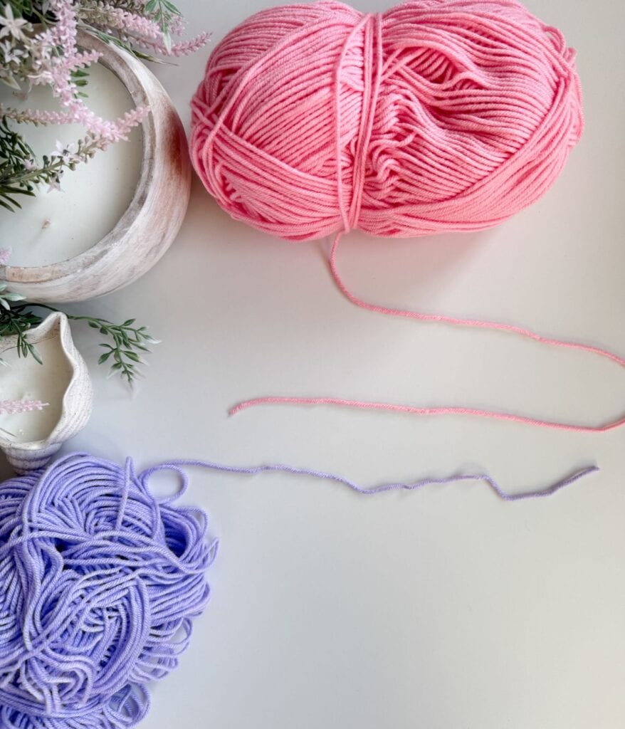 A ball of pink yarn and a ball of purple yarn sit on a white surface, with decorative plants and a white bowl nearby—perfect for practicing crochet techniques such as how to join new yarn in crochet projects.