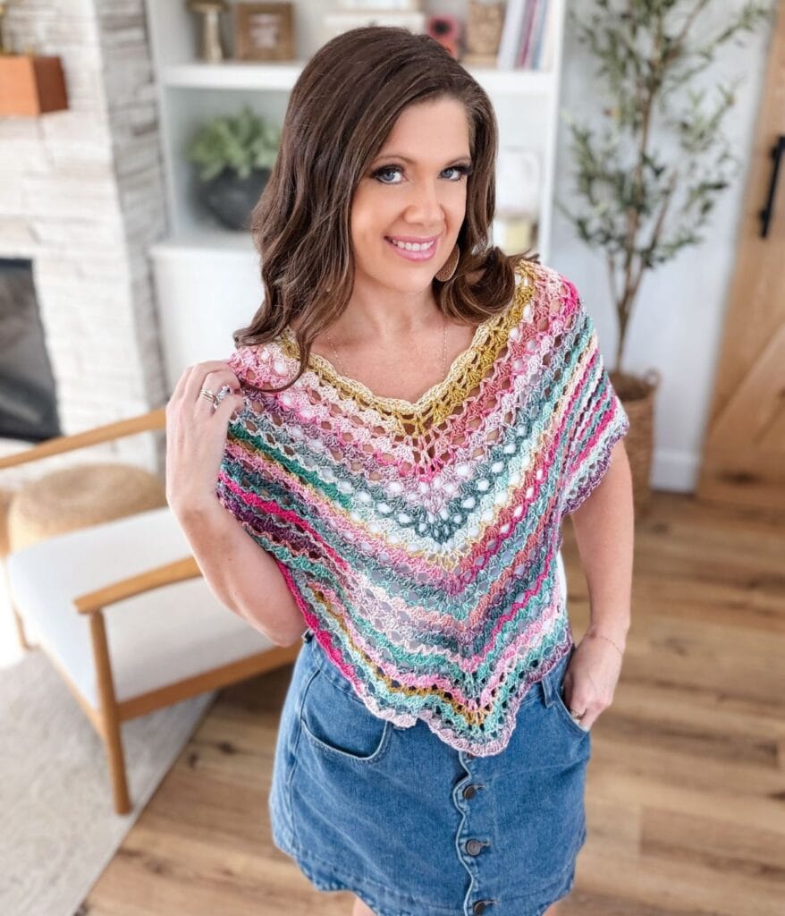 A woman wearing a colorful crochet top, inspired by a free crochet poncho pattern, and a denim skirt stands in a living room with wooden floors and modern decor.