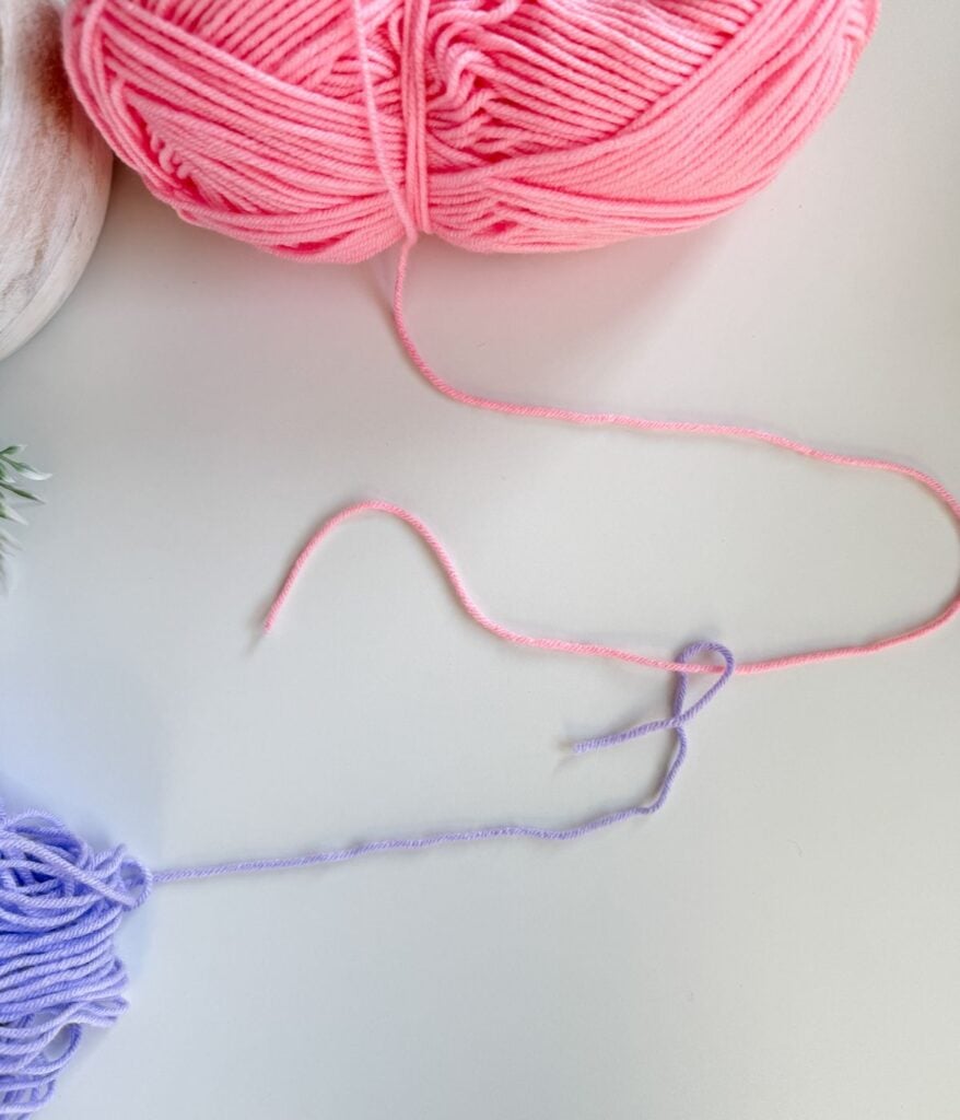 A pink and a purple yarn ball are shown on a white surface, with their ends tied together in a simple knot, demonstrating an easy crochet technique for how to join yarn.