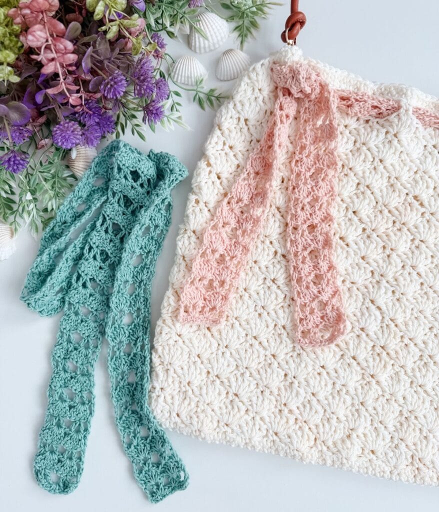 Crocheted cream bag with a pink ribbon, a separate teal crocheted ribbon, and a floral arrangement with seashells in the background—this charming accessory doubles as an elegant Beach Bag. Crafted from a Free Crochet Pattern, it brings coastal style to your everyday outings.