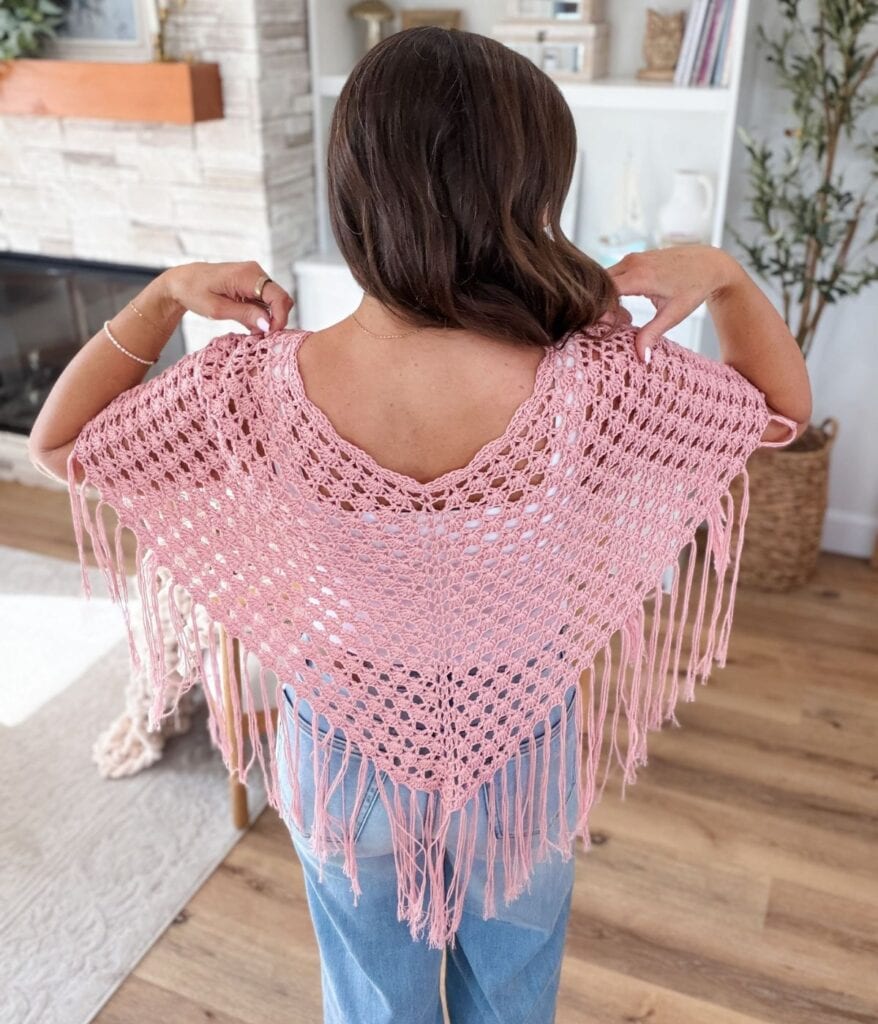 A person wearing a pink crocheted shawl with fringes stands indoors, facing away and holding the shawl open to display its design, perfect inspiration if you're searching for a free crochet poncho pattern.