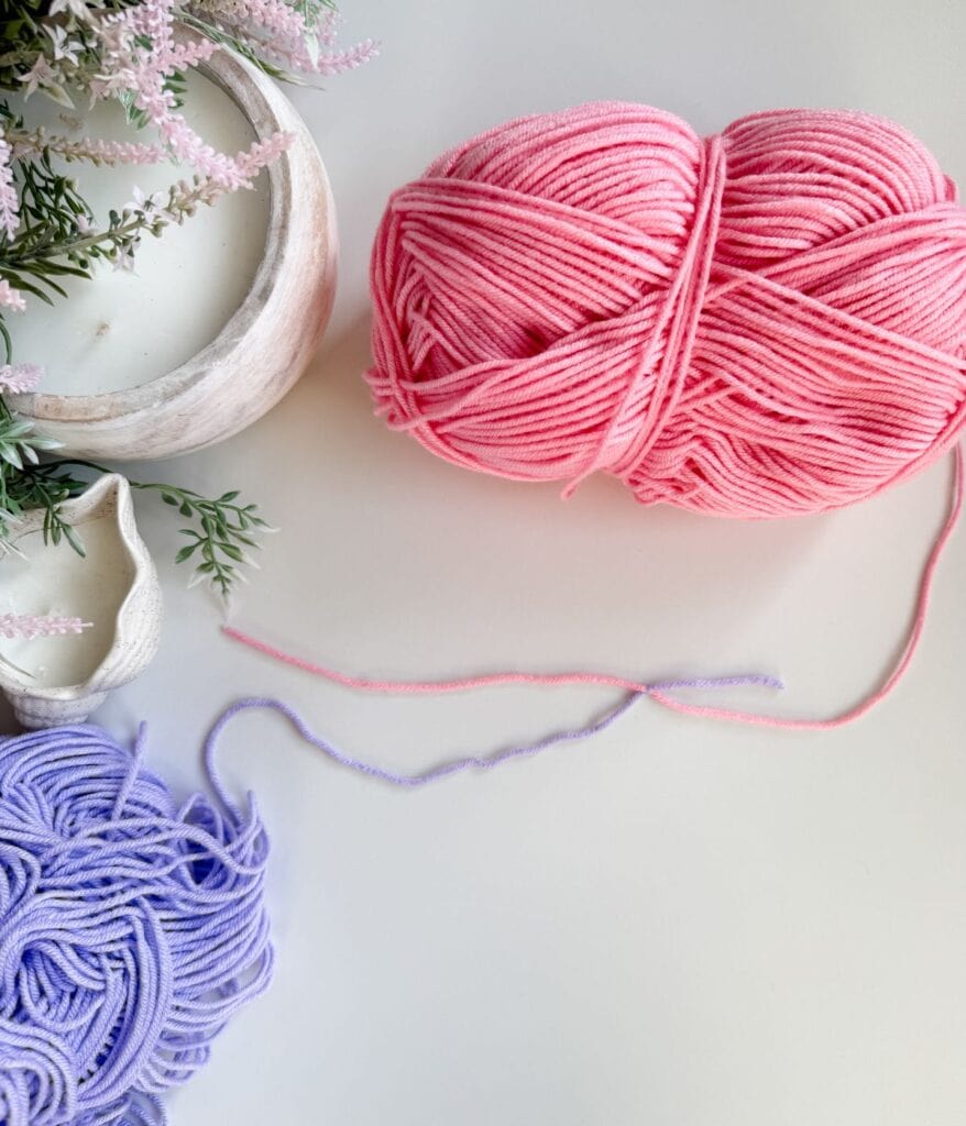 Pink and purple yarn balls on a white surface next to a potted plant, a white bowl, and a seashell—perfect for exploring new crochet techniques or learning how to join yarn as you start your next creative project.