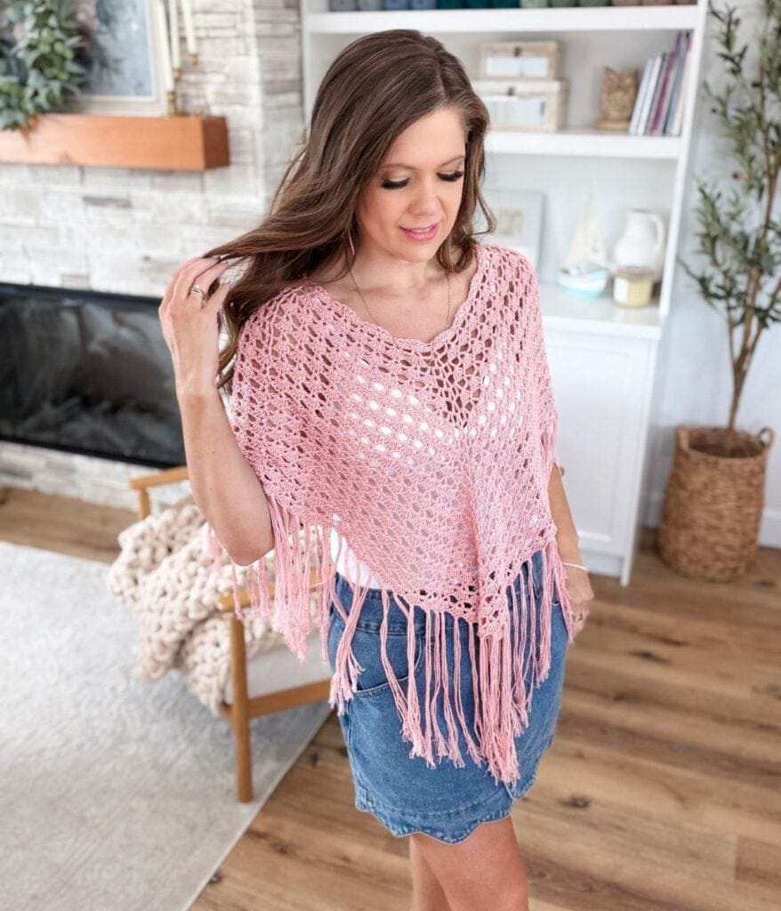 Woman wearing a pink crocheted fringed poncho—made from a free crochet poncho pattern—and a blue denim skirt stands indoors on a wooden floor near a fireplace and white shelves.