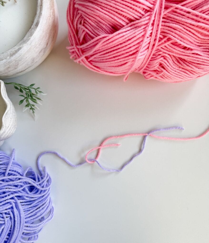 Pink and purple yarn balls sit on a white surface, with loose strands stretching between them—perfect for practicing crochet techniques or learning how to join new yarn in crochet projects. A potted plant is partially visible on the left.