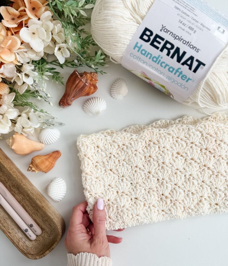A hand holds a textured white crochet piece next to white yarn, crochet hooks, seashells, and artificial flowers on a white surface, hinting at the start of a stunning Crochet Beach Bag Pattern—perfect for your next seaside adventure.
