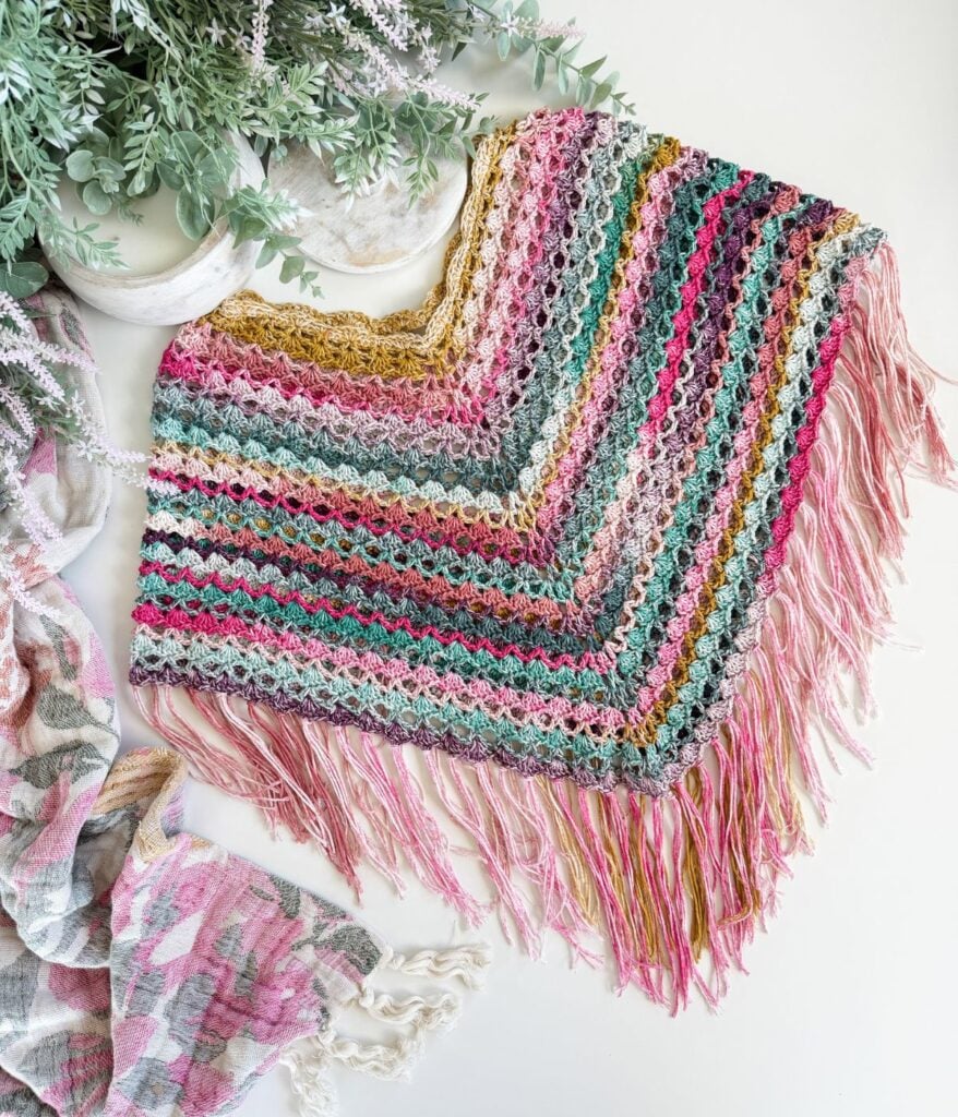 A colorful crochet poncho with pink, green, gold, and white stripes, featuring fringe along the edges, is displayed beside a green plant and a patterned cloth—a lovely creation inspired by a free crochet poncho pattern.