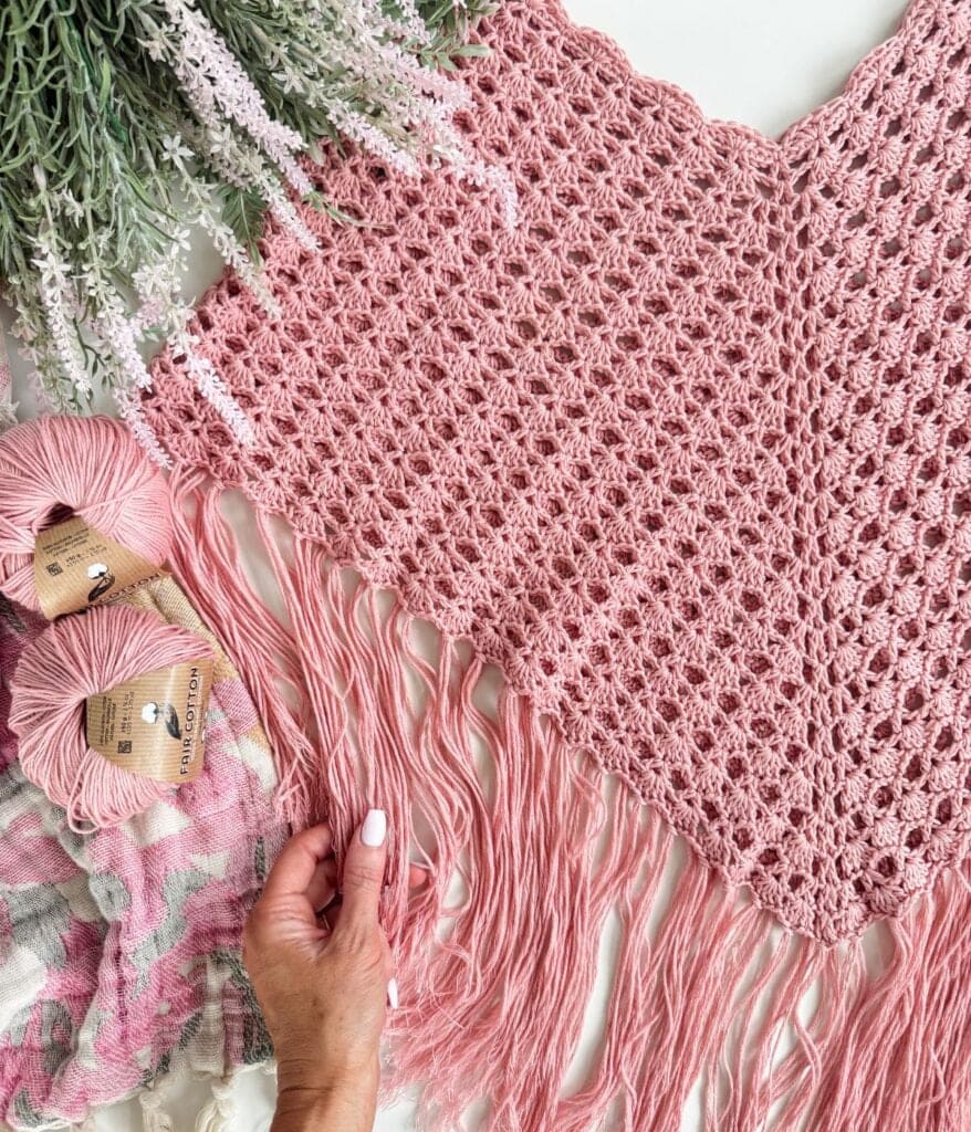 Pink crocheted fabric with a fringe edge is displayed alongside pink yarn balls, a hand gently touching the yarn, and lavender flowers arranged above—perfect inspiration for trying out a free crochet poncho pattern.