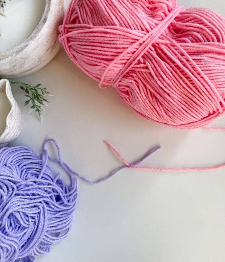 A ball of pink yarn and a loose pile of purple yarn rest on a white surface, ready to demonstrate how to join new yarn in crochet, with some green leaves and ceramic dishes partially visible in the corners.